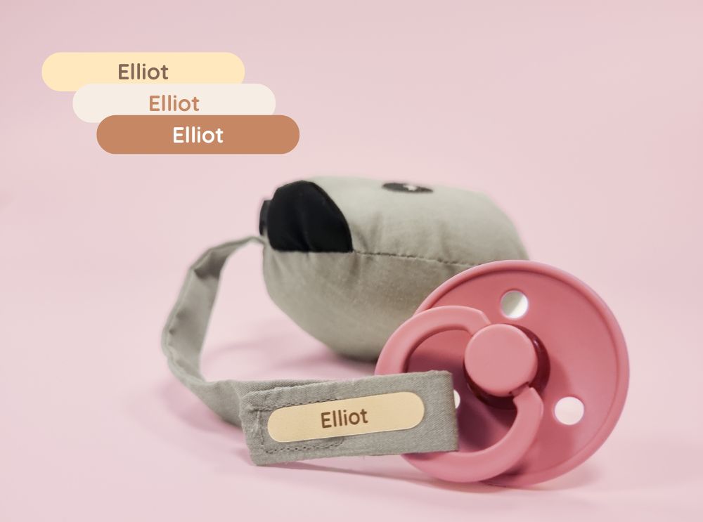 Roze fopspeen met een gepersonaliseerd naamsticker voor elliot aan een stoffen band.