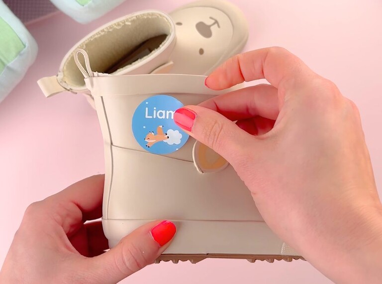 Rond blauw naamlabel met vosje wordt op een beige kinderschoen geplakt