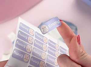 Hand holding a purple personalized name label with a bunny design and the name Emmy, peeled from a sticker sheet