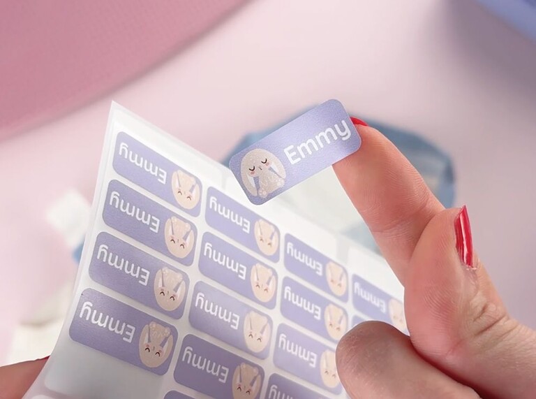 Hand holding a purple personalized name label with a bunny design and the name Emmy, peeled from a sticker sheet