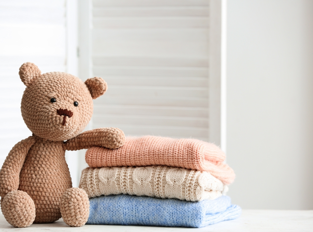 Knitted teddy bear sitting next to a stack of folded baby clothes in soft pastel colors