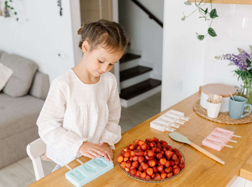 Meisje bereidt aardbeien voor aan de keukentafel met ijsvormpjes in de buurt – klaar om zelfgemaakte bevroren lekkernijen te maken.