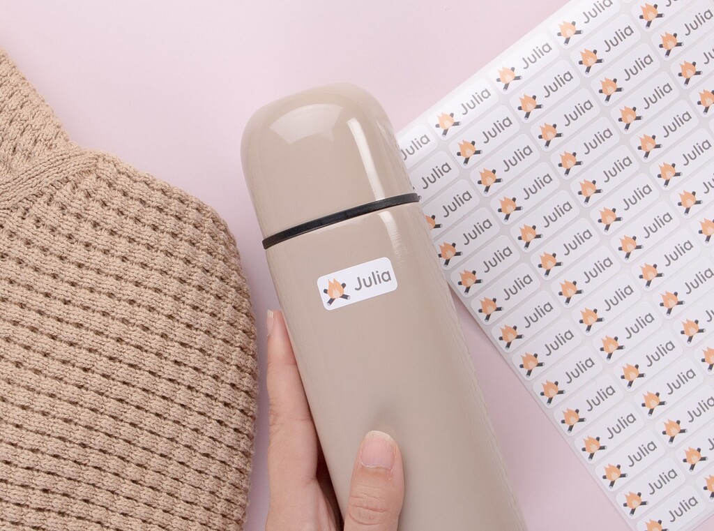 Extra kleren op het kinderdagverblijf 1 Hand die een beige drinkfles vasthoudt met een wit naametiket met een kampvuur en de naam julia, naast een bijpassend stickervel.