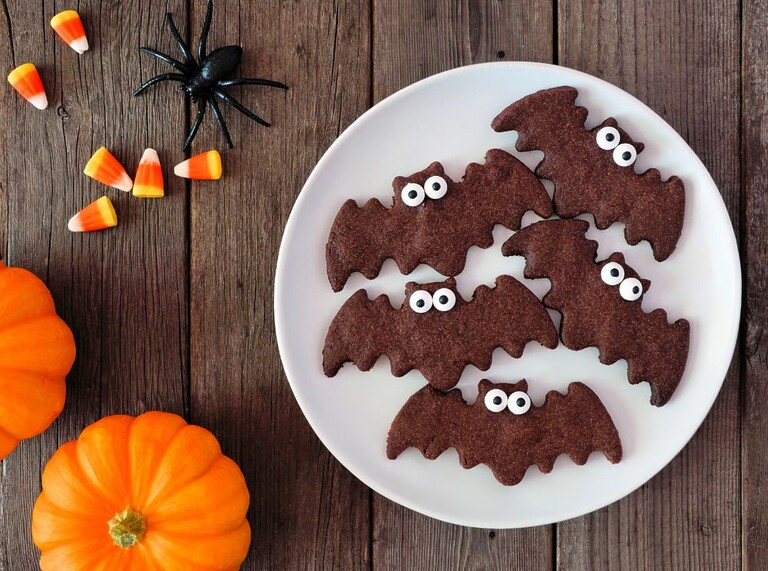 Plate of fun bat-shaped Halloween cookies with candy eyes, surrounded by pumpkins and candy corn — a perfect kid-friendly Halloween treat.