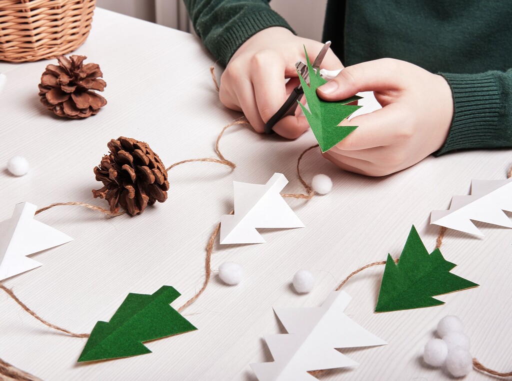 Kind dat een kerst­slinger maakt met papieren boompjes, dennenappels en touw voor een leuke kerstknutsel.