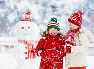 Two kids standing in front of a snowman, warmly dressed and holding cups of hot cocoa while snow is falling.