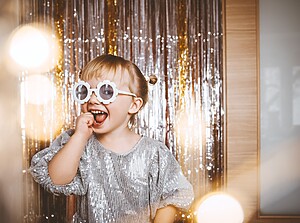 Happy child wearing flower-shaped sunglasses and a sparkly silver outfit in front of a glitter backdrop