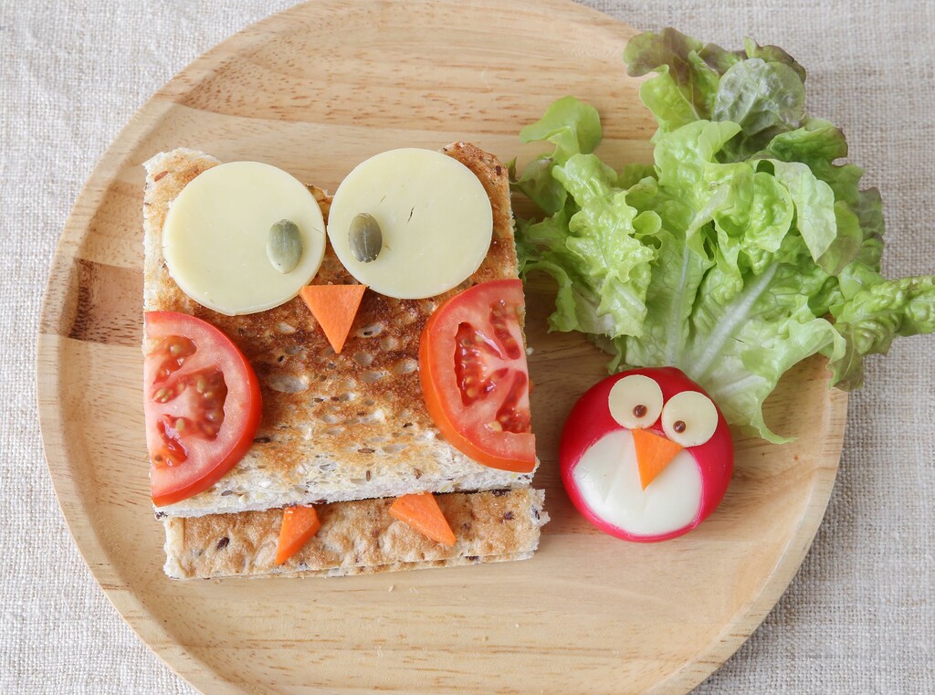 Uilvormige kindersnack van toast, kaas, tomaat en wortel met een schattig ei-kuikentje op een houten bord.