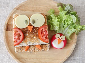 Uilvormige kindersnack van toast, kaas, tomaat en wortel met een schattig ei-kuikentje op een houten bord.