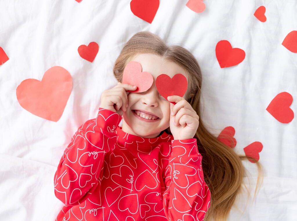 Smiling child holding red paper hearts over their eyes, lying on a white background with heart decorations.