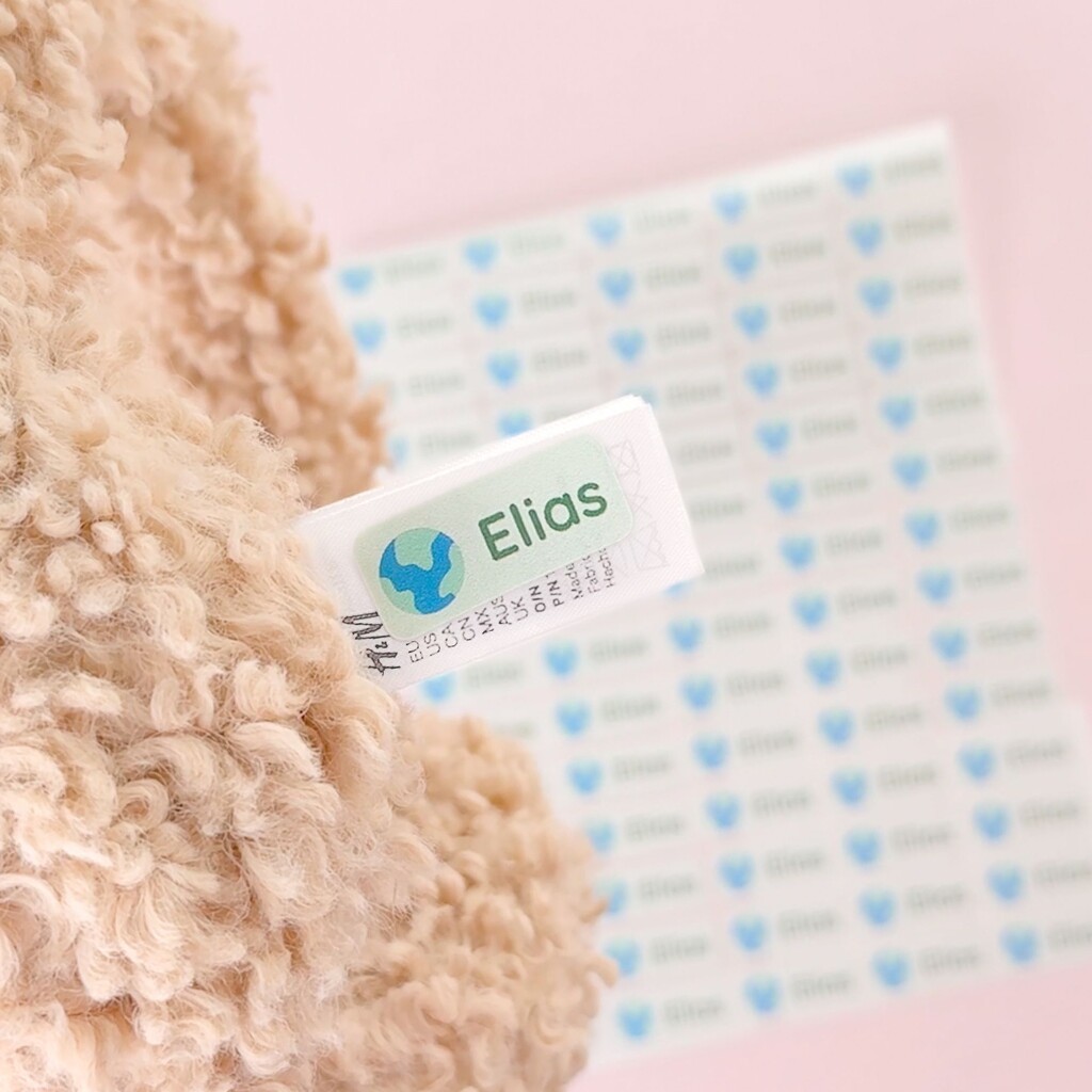 Close-up van een gepersonaliseerd naamsticker met de naam elias en een wereldbol-icoon op een kledinglabel, met een bijpassend stickervel op de achtergrond.